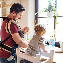 Ein Vater hat ein Baby in der Trage und steht an der Spüle. Ein weiteres Kleinkind steht neben ihm auf einem Hocker an der Spüle.