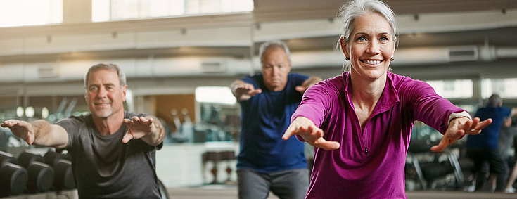 Eine Frau und zwei Männer machen gemeinsam Sportübungen in einem Fitnessraum.