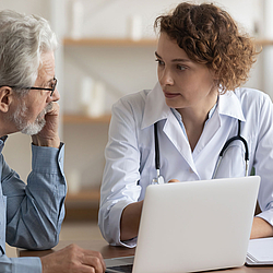 Eine Ärztin spricht mit einem älteren Patienten. Sie zeigt ihm etwas auf ihrem Laptop.