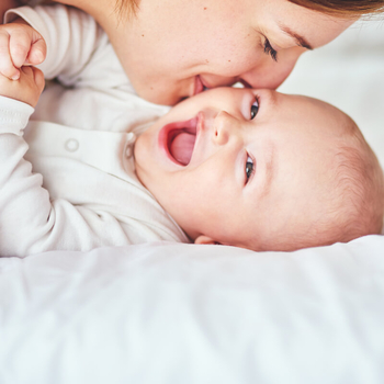 Ein Baby in einem weißen Body liegt auf einer Matratze. Es lacht fröhlich in die Kamera. Seine Mutter lächelt und gibt ihm einen Kuss auf die Wange.