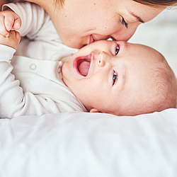 Ein Baby in einem weißen Body liegt auf einer Matratze und lächelt. Seine Mutter gibt ihm einen Kuss auf die Wange.