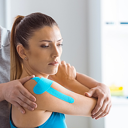 	Ein Physiotherapeut führt bei einer Frau eine Schulterbehandlung durch. Die Patientin hat Kinesio-Tape auf Arm und Schulter.