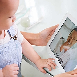 Ein Baby in einer blauen Latzhose sitzt auf dem Schoß seiner Mutter, beide schauen auf ein weißes Tablet. Auf dem Bildschirm sieht man einen jungen Arzt in einem weißen Kittel.