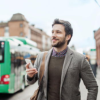 Ein Mann steht lächelt in der Stadt und hält sein Smartphone. Im Hintergrund steht ein Bus.