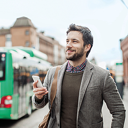 Ein Mann steht lächelt in der Stadt und hält sein Smartphone. Im Hintergrund steht ein Bus.