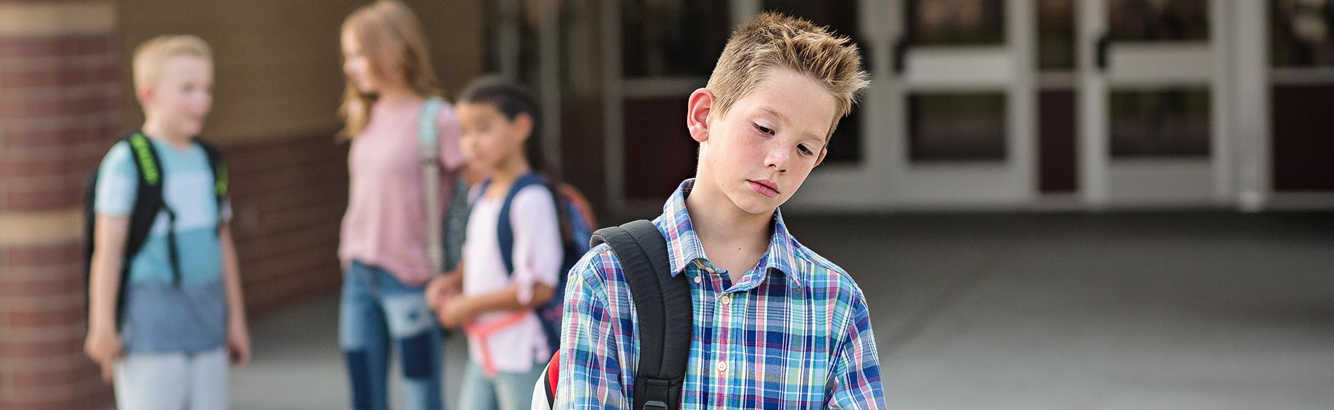 Schüler steht mit gesenktem Blick auf dem Schulhof, während andere Kinder im Hintergrund zusammenstehen.
