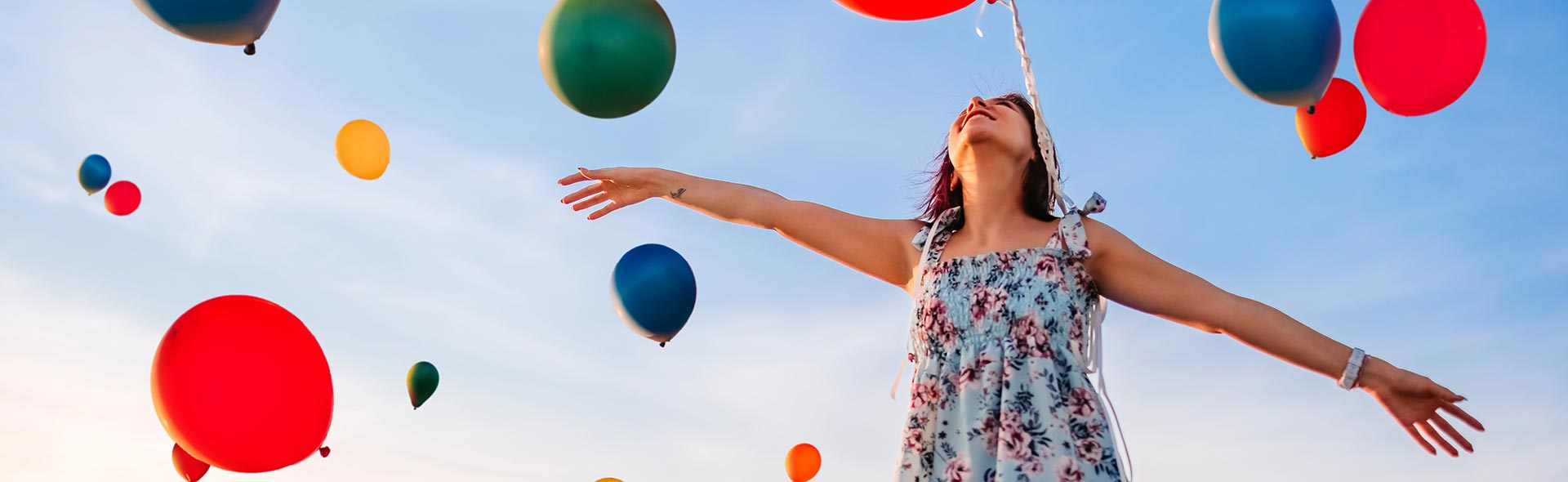 Person mit ausgestreckten Armen unter bunten Luftballons vor blauem Himmel.