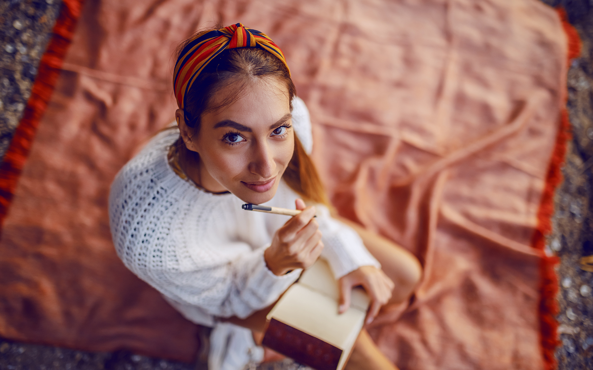 Eine junge Frau mit langen Haaren und einem bunten Tuch im Haar, sitzt auf einer roten Decke. Sie trägt einen weißen Pullover, auf ihrem Schoß liegt ein Notizbuch. In ihrer Hand hält sie einen Stift. Sie schaut lächelnd nach oben in die Kamera.